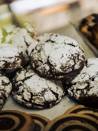 Close-up of chocolate crinkle cookies dusted with powdered sugar on a bakery display tray with spiral pinwheel cookies in the foreground