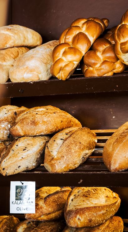 Warm golden braided challah and rustic artisan loaves, including Kalamata olive sourdough, stacked on wooden bakery shelves