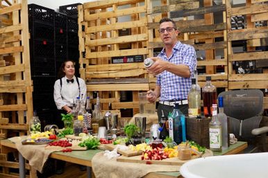 Man in blue checked shirt shaking a cocktail shaker at a rustic bar setup with bottles, fresh fruit, herbs and glassware while a server in white shirt and bow tie watches in front of stacked wooden pallets.
