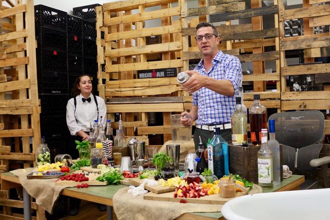 Man in blue checked shirt shaking a cocktail shaker at a rustic bar setup with bottles, fresh fruit, herbs and glassware while a server in white shirt and bow tie watches in front of stacked wooden pallets.