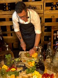 Mixologist in an apron slicing limes at a rustic wooden bar prep station, surrounded by oranges, lemons, berries, ginger, fresh herbs, jars and bottles for cocktail garnishes.