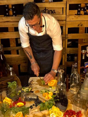 Mixologist in an apron slicing limes at a rustic wooden bar prep station, surrounded by oranges, lemons, berries, ginger, fresh herbs, jars and bottles for cocktail garnishes.