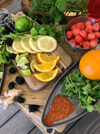 Colorful citrus and herbs on a rustic wooden board: lemon and orange slices, lime wedges, watermelon balls, blackberries, ginger, cilantro and chili powder — fresh garnish prep.