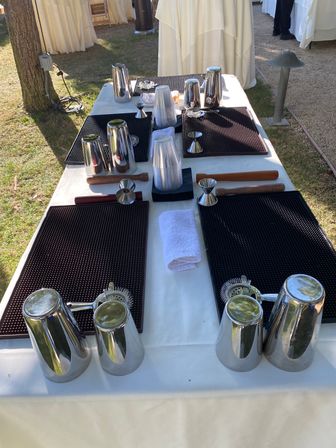 Outdoor cocktail station on a lawn with stainless steel shakers, jiggers, strainers, muddlers, plastic cups and black bar mats on a white tablecloth.