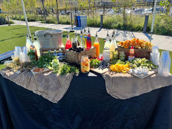 Sunny park drink station with metal ice tub, colorful bottled juices, stacked plastic cups and platters of sliced citrus, berries and fresh herbs for cocktails and mocktails.