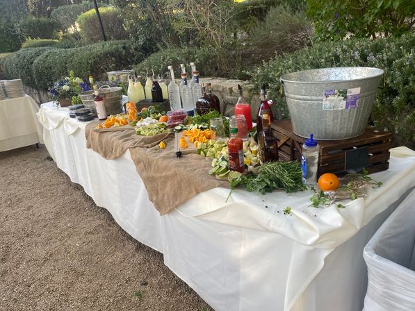 Outdoor garden cocktail station on a white-tablecloth and burlap-covered table with metal tubs and crates, assorted liquor bottles and mixers, jars and bottles of juices, bowls of sliced citrus, cucumber and fresh herbs, and garnish platters for a party.
