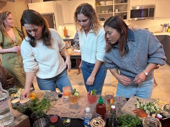 Three friends gathered at a modern kitchen island making cocktails, glasses garnished with citrus and herbs, cutting board of lime wedges, jars, mixers and fresh greens scattered on the counter during a casual home gathering.
