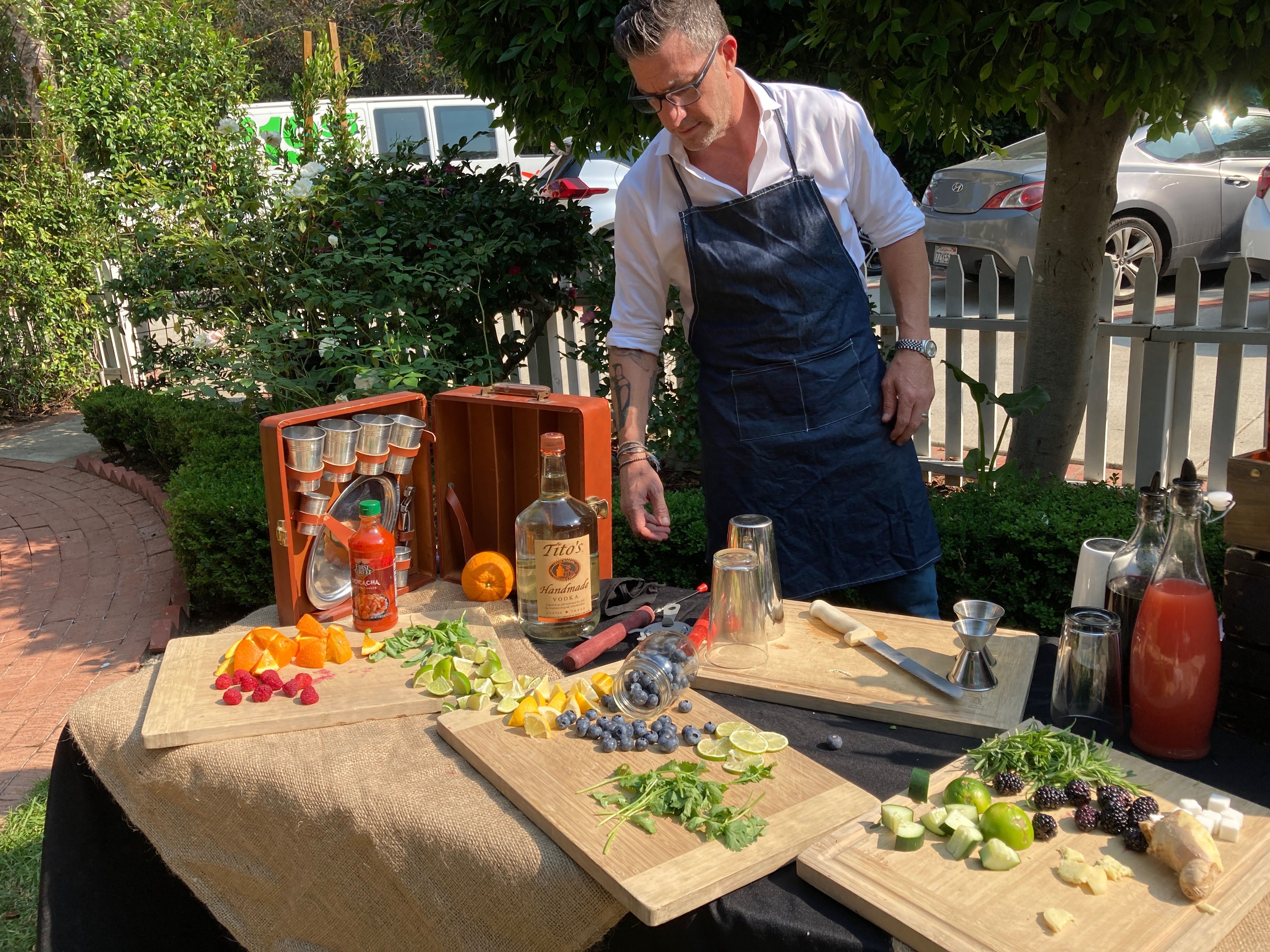Sunny outdoor garden cocktail station with an apron-clad mixologist arranging sliced oranges, limes, berries, cucumber, ginger and fresh herbs on wooden boards, surrounded by bottles, shakers, a jigger and other bartending tools on a burlap-covered table.
