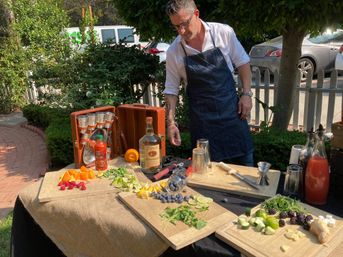Sunny outdoor garden cocktail station with an apron-clad mixologist arranging sliced oranges, limes, berries, cucumber, ginger and fresh herbs on wooden boards, surrounded by bottles, shakers, a jigger and other bartending tools on a burlap-covered table.