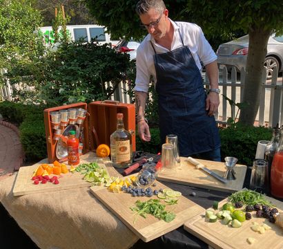 Sunny outdoor garden cocktail station with an apron-clad mixologist arranging sliced oranges, limes, berries, cucumber, ginger and fresh herbs on wooden boards, surrounded by bottles, shakers, a jigger and other bartending tools on a burlap-covered table.