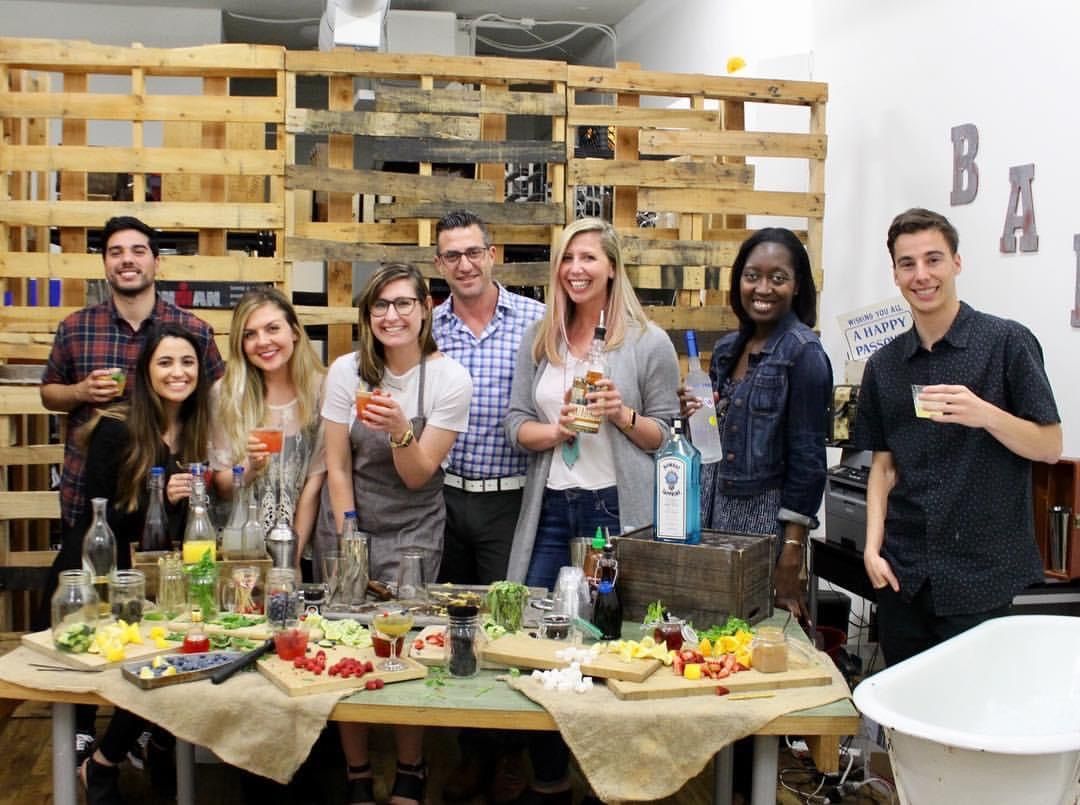 Smiling group of friends holding colorful cocktails around a table of fresh fruit, syrups and liquor bottles in a casual indoor mixology workshop with a wooden pallet backdrop.