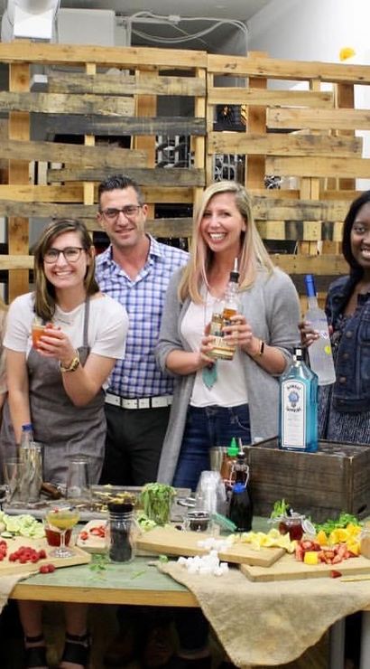 Smiling group of friends holding colorful cocktails around a table of fresh fruit, syrups and liquor bottles in a casual indoor mixology workshop with a wooden pallet backdrop.