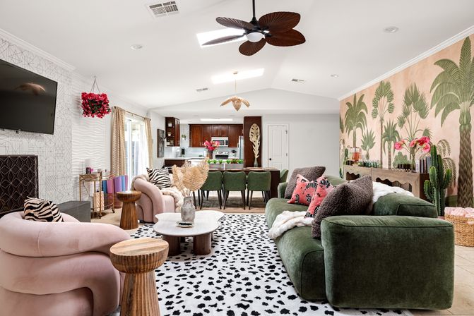 Open-plan modern boho living room and kitchen with green velvet sofa, blush pink curved chairs, black-and-white spotted rug, large tropical palm wall mural, wooden side tables, hanging plant, and dark wood kitchen cabinets.