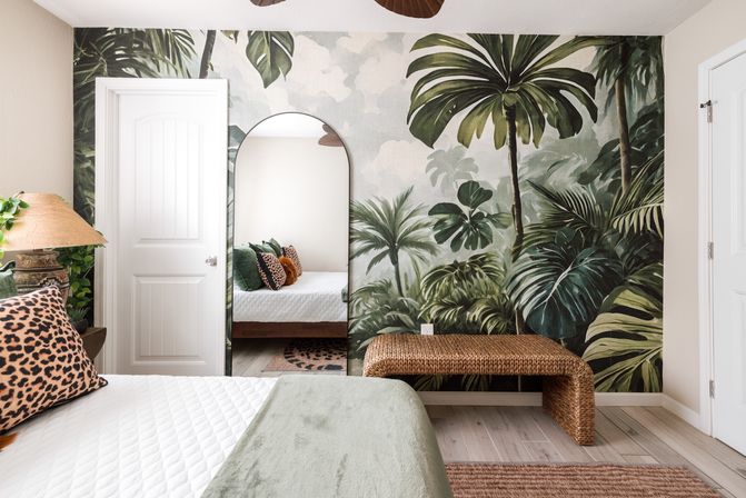 Tropical-themed bedroom with oversized palm and monstera wallpaper mural, arched full-length mirror reflecting a bed with leopard-print accent pillows, woven rattan bench, lamp and green throw — nature-inspired, airy decor.