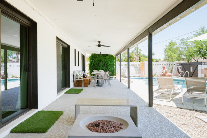 Sunny covered backyard patio with ceiling fans, outdoor dining set, bench and fire pit overlooking lounge chairs, swimming pool and green hedge