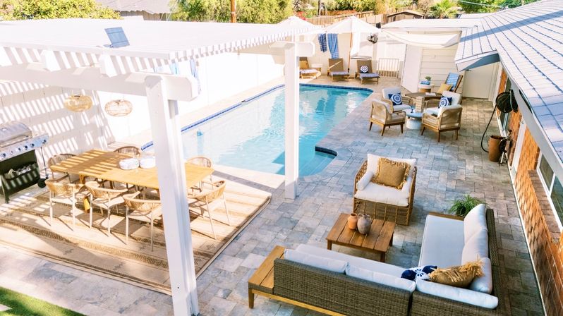 Sun-drenched suburban backyard with rectangular swimming pool, pergola-shaded dining table and grill, and wicker lounge seating arranged on a stone-paver patio.