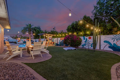 Cozy backyard patio at sunset with lit swimming pool, Adirondack chairs around a fire pit, string lights, pergola bar, colorful mural fence, palm trees and cactus