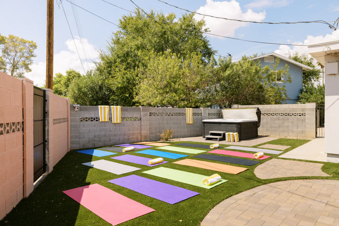 Sunny suburban backyard set up for outdoor yoga with colorful mats and rolled towels on artificial turf, hot tub by a cinderblock wall