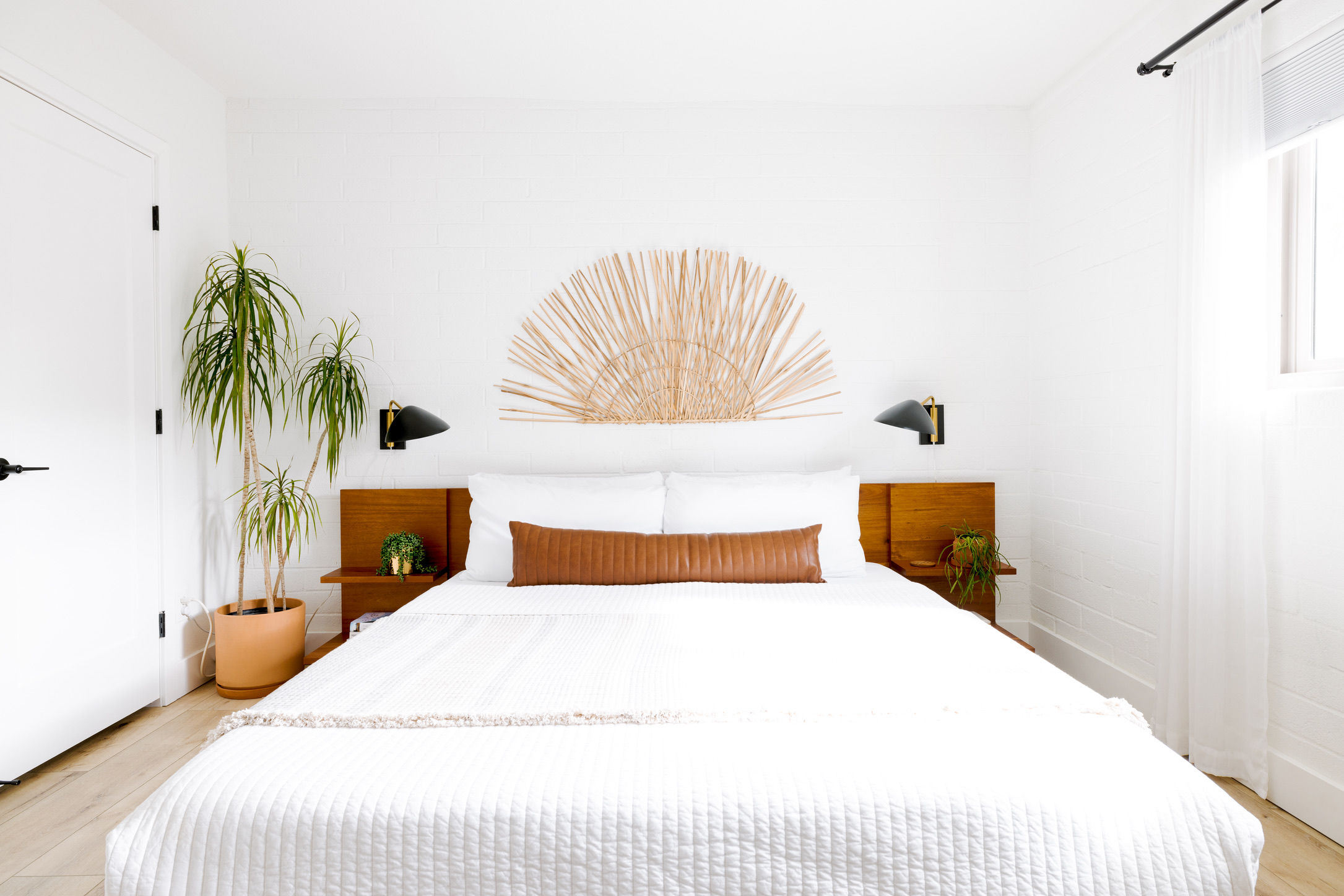 Bright modern minimalist bedroom with white quilted bedding, wooden headboard, tan leather lumbar pillow, boho sunburst wall art, potted palm, black wall sconces and sheer curtains letting in natural light