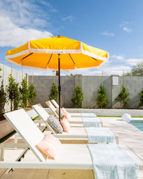 Sunny backyard poolside scene with a cheery yellow umbrella shading white lounge chairs dressed with pastel pillows and light-blue towels by a cinderblock privacy wall.