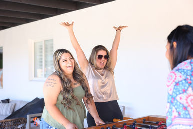 Three people playing foosball on a sunlit covered patio — two in the foreground laugh and cheer, one raising her arms in a celebratory pose.