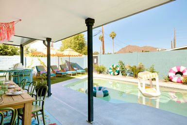 Sunny desert backyard with covered patio and outdoor dining, turquoise pool with striped inflatables, lounge chairs under yellow umbrellas, palm trees and distant rocky hill