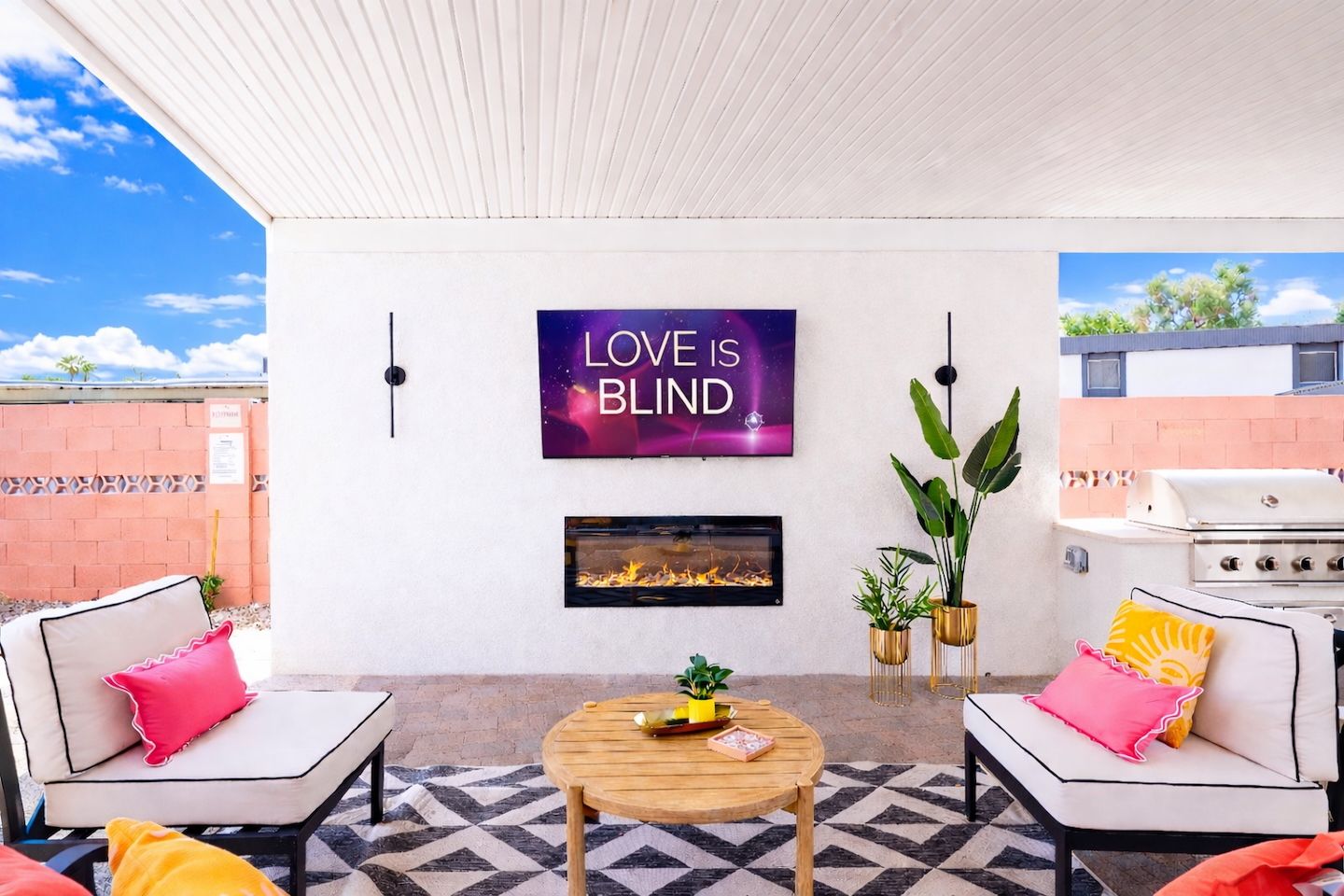 Sunny covered backyard patio and outdoor living area with wall-mounted TV reading "LOVE IS BLIND" above a linear fireplace, cushioned lounge chairs with pink and yellow accent pillows, round wooden coffee table on a geometric rug, potted plants and stainless steel grill.