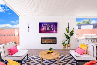 Sunny covered backyard patio and outdoor living area with wall-mounted TV reading "LOVE IS BLIND" above a linear fireplace, cushioned lounge chairs with pink and yellow accent pillows, round wooden coffee table on a geometric rug, potted plants and stainless steel grill.