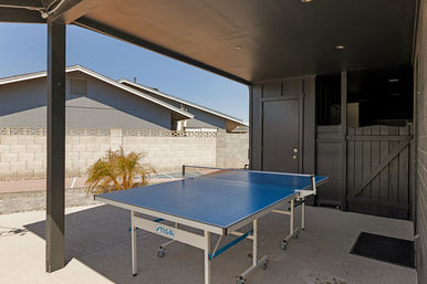 Covered backyard patio with game-ready blue outdoor ping pong table on wheels, concrete floor, gray cinderblock privacy wall, small palm, and neighboring single-story roofs under a clear blue sky.