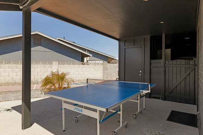 Covered backyard patio with game-ready blue outdoor ping pong table on wheels, concrete floor, gray cinderblock privacy wall, small palm, and neighboring single-story roofs under a clear blue sky.