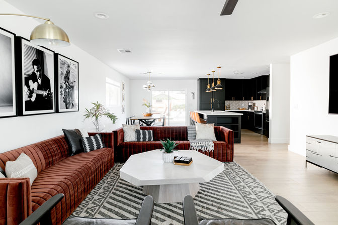Sunny open-concept modern living room and kitchen featuring a rust velvet sectional, geometric gray rug, hexagonal white coffee table, black kitchen island with brass pendant lights, and sliding glass door letting in natural light.