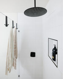 Spa-like modern shower with matte black rain showerhead and fixtures, white tiled walls, recessed niche holding shampoo bottle, and light striped towels on glass hooks.