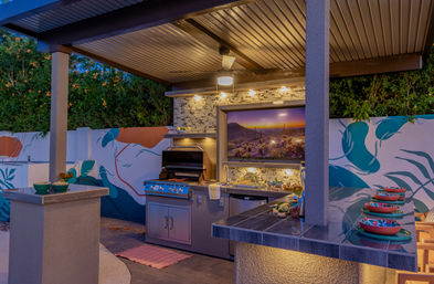 Backyard covered outdoor kitchen at dusk with a built-in stainless steel grill, stone-tiled backsplash and wall-mounted TV showing a desert sunset, colorful botanical mural on the perimeter wall and a tiled bar with stacked bowls.