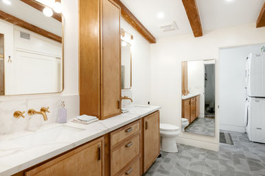 Bright modern farmhouse-style bathroom with long oak vanity and marble countertop, brass wall-mounted faucets, exposed wood beams, patterned gray tile floor, full-length mirror and stacked washer-dryer in an adjoining laundry nook.