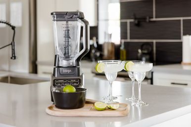 Countertop blender on a modern kitchen island with three lime‑garnished margarita glasses and halved limes on a wooden board, ready to serve.