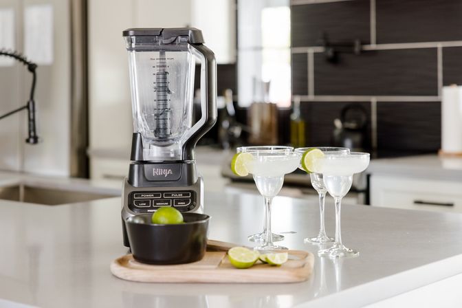Countertop blender on a modern kitchen island with three lime‑garnished margarita glasses and halved limes on a wooden board, ready to serve.