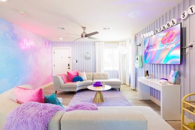 Playful pastel living room in a modern apartment: lavender and pink gradient wall with neon 'Lover' sign, curved light-gray sectional with bright pillows, plush purple rug and throws, brass coffee table, wall-mounted TV showing colorful graphics, mint guitar and striped wallpaper.