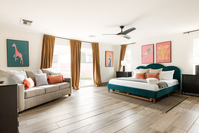 Sunlit modern master bedroom interior with teal upholstered bed, coral accent pillows, beige sofa with striped cushions, golden floor-to-ceiling curtains, wood-look tile flooring, black ceiling fan and playful animal artwork on the walls.