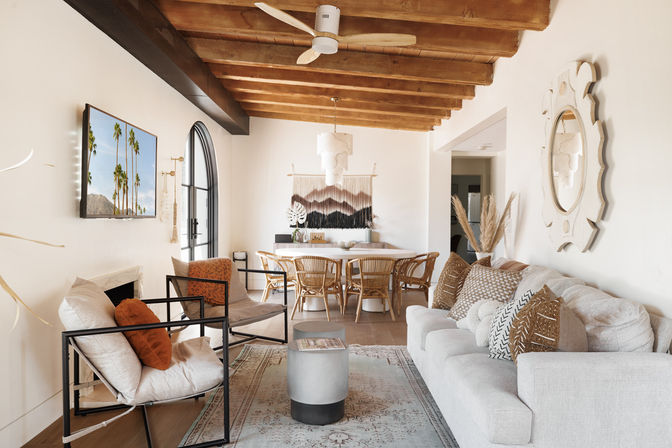 Sunlit coastal-boho open-plan living and dining room with exposed wooden beam ceiling, arched black-framed window, rattan dining chairs, neutral sofa with patterned pillows, woven wall hanging and pampas grass.