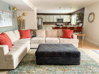 Open-concept living room with beige L-shaped sectional, coral textured throw pillows, oversized black ottoman on a colorful area rug, and a white kitchen with patterned backsplash and pendant lights.