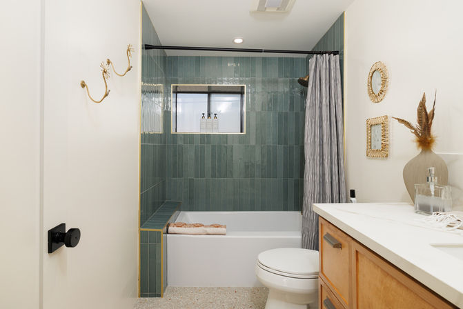 Modern residential bathroom with vertical green subway-tiled shower and built-in bench, white tub and toilet, gray shower curtain, wooden vanity with white countertop, terrazzo floor, gold wall hooks and woven decor