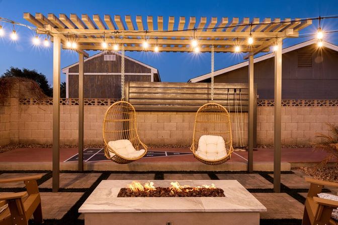 Cozy backyard patio at dusk with a pergola draped in string lights, two hanging rattan chairs with cushions above a stone fire pit and a shuffleboard court in the background.