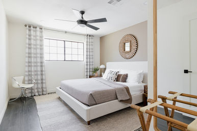 Bright modern bedroom with neutral tones — upholstered platform bed with gray bedding and patterned pillows, round woven mirror, large window with plaid curtains, ceiling fan, clear acrylic chair and wooden luggage rack.