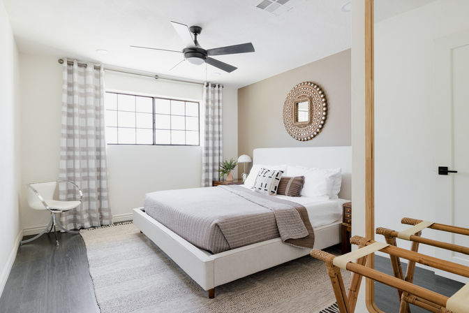 Bright modern bedroom with neutral tones — upholstered platform bed with gray bedding and patterned pillows, round woven mirror, large window with plaid curtains, ceiling fan, clear acrylic chair and wooden luggage rack.