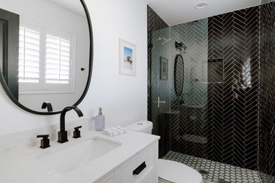 Modern white bathroom with marble-look vanity, matte black faucet and round mirror, shuttered window, glass-enclosed shower featuring glossy black herringbone wall tile and hexagon floor tile.