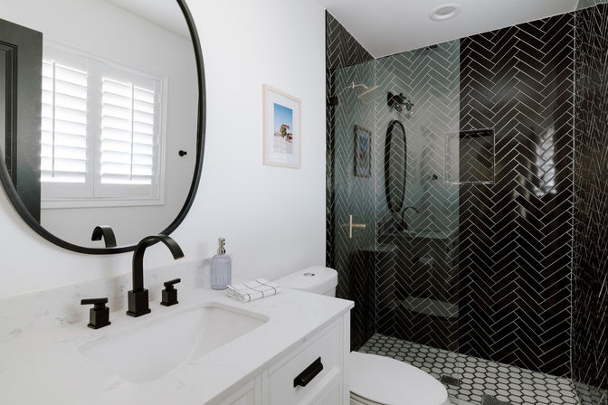 Modern white bathroom with marble-look vanity, matte black faucet and round mirror, shuttered window, glass-enclosed shower featuring glossy black herringbone wall tile and hexagon floor tile.