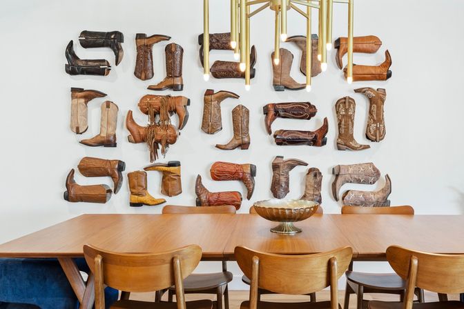 Midcentury wooden dining table and chairs under a brass pendant light, against a white wall arranged with mounted Western cowboy boot halves in a grid and a decorative bowl on the table.