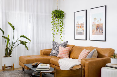 Cozy sunlit living room with a tan leather sectional, patterned throw pillows and blanket, hanging pothos and large floor plant, black-framed wall art, and a low black coffee table with books.