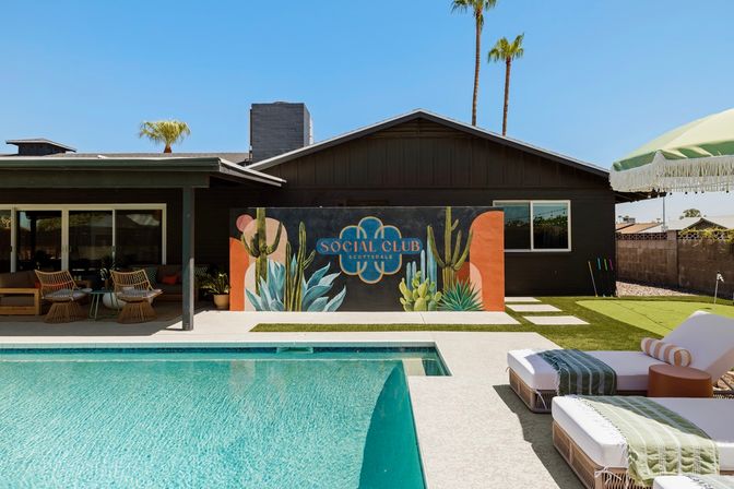 Sunlit Southwestern backyard with a turquoise swimming pool, cushioned lounge chairs, shaded patio seating and a vibrant cactus mural on a dark mid-century house wall, palm trees rising in the background.