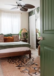 Sunlit bedroom seen through an open green door, featuring a wooden bed with green throw and leopard-print pillows, rust fur accent pillow, palm-leaf wallpaper, wicker lamp, sculptural ceiling fan and a bold leopard-face area rug.